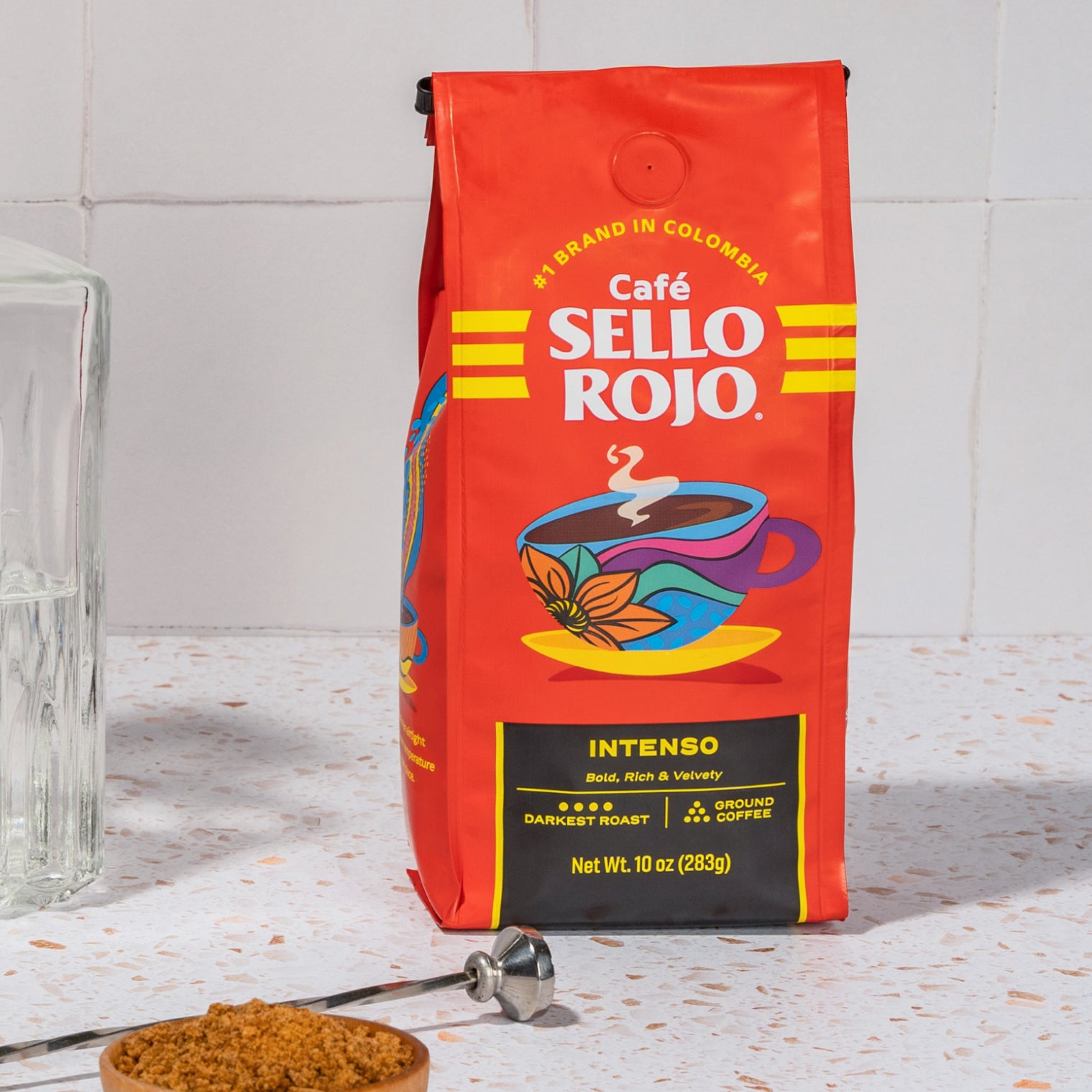 Red coffee bag labeled 'Café Sello Rojo' on a light surface with a glass and small bowl of coffee grounds.