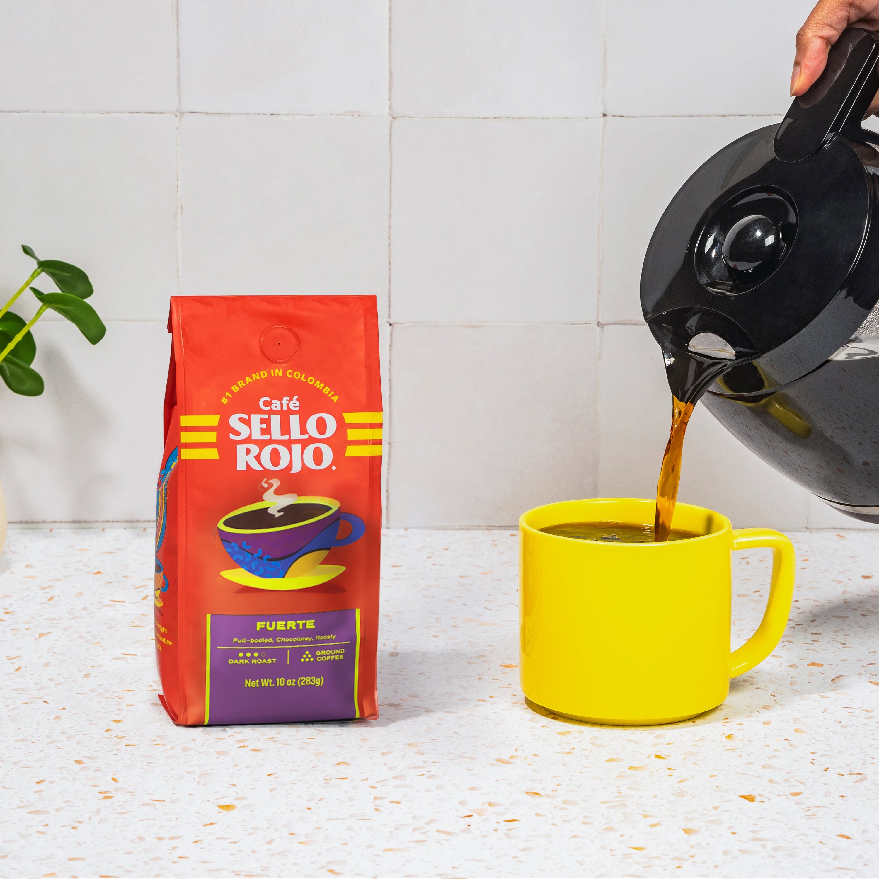 A person pours coffee from a black electric kettle into a bright yellow mug next to a red bag of Cafe Sello Rojo, set on a white countertop